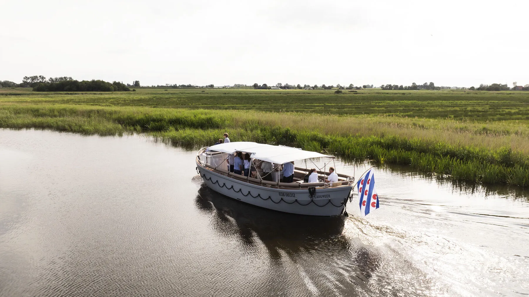 De sloep met wapperende Friese vlag vaart door een smalle vaart tussen de weilanden